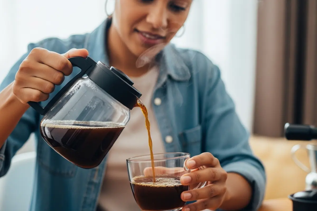 Close em uma mulher de camisa jeans sorrindo enquanto serve café de uma jarra de vidro em uma xícara transparente.