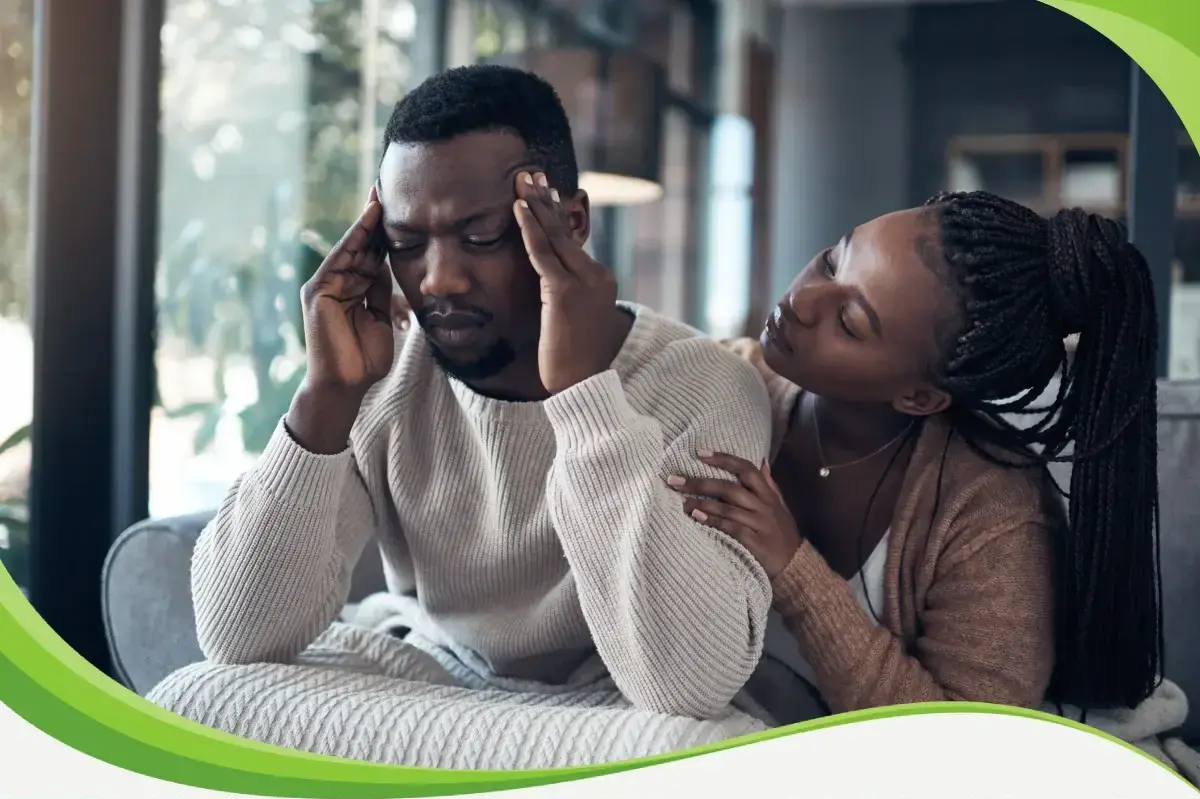 Mulher consolando companheiro no sofá, abraçada nele. Ele está tocando as têmporas, com feições de dor.
