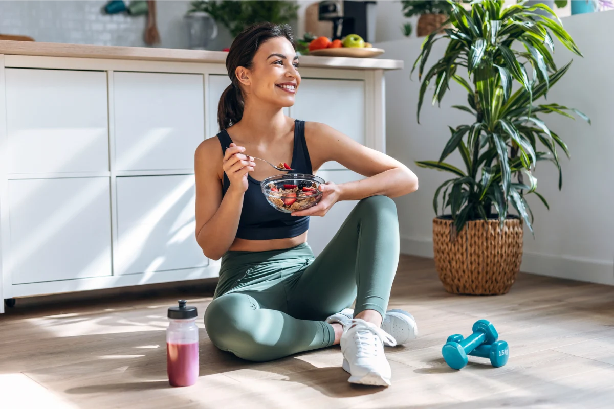 Mulher com roupas de academia, sentada no chão, comendo frutas de um bowl e sorrindo.