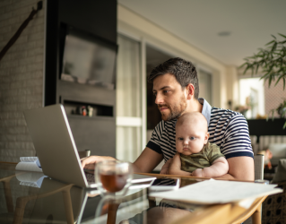 Pai sentado à mesa segurando um bebê no colo enquanto olha atentamente para a tela de um notebook, pesquisando sobre a dosagem correta de medicação.